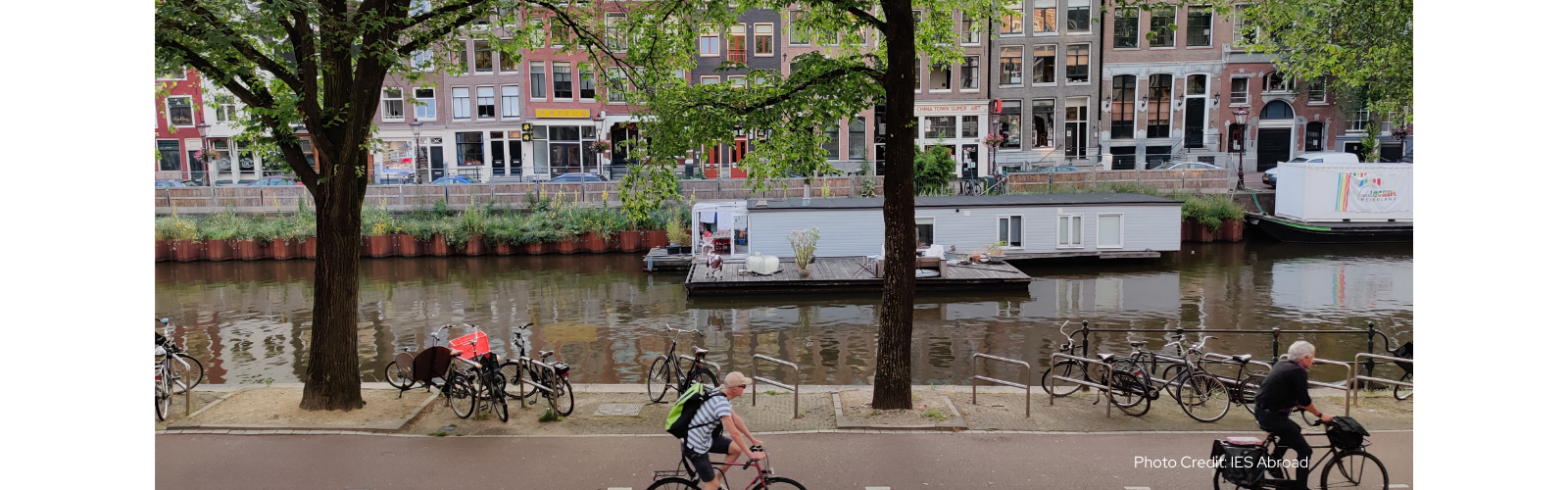 People pass by on bikes in front of a river with two houseboats, and there are several tall narrow buildings in the background. 